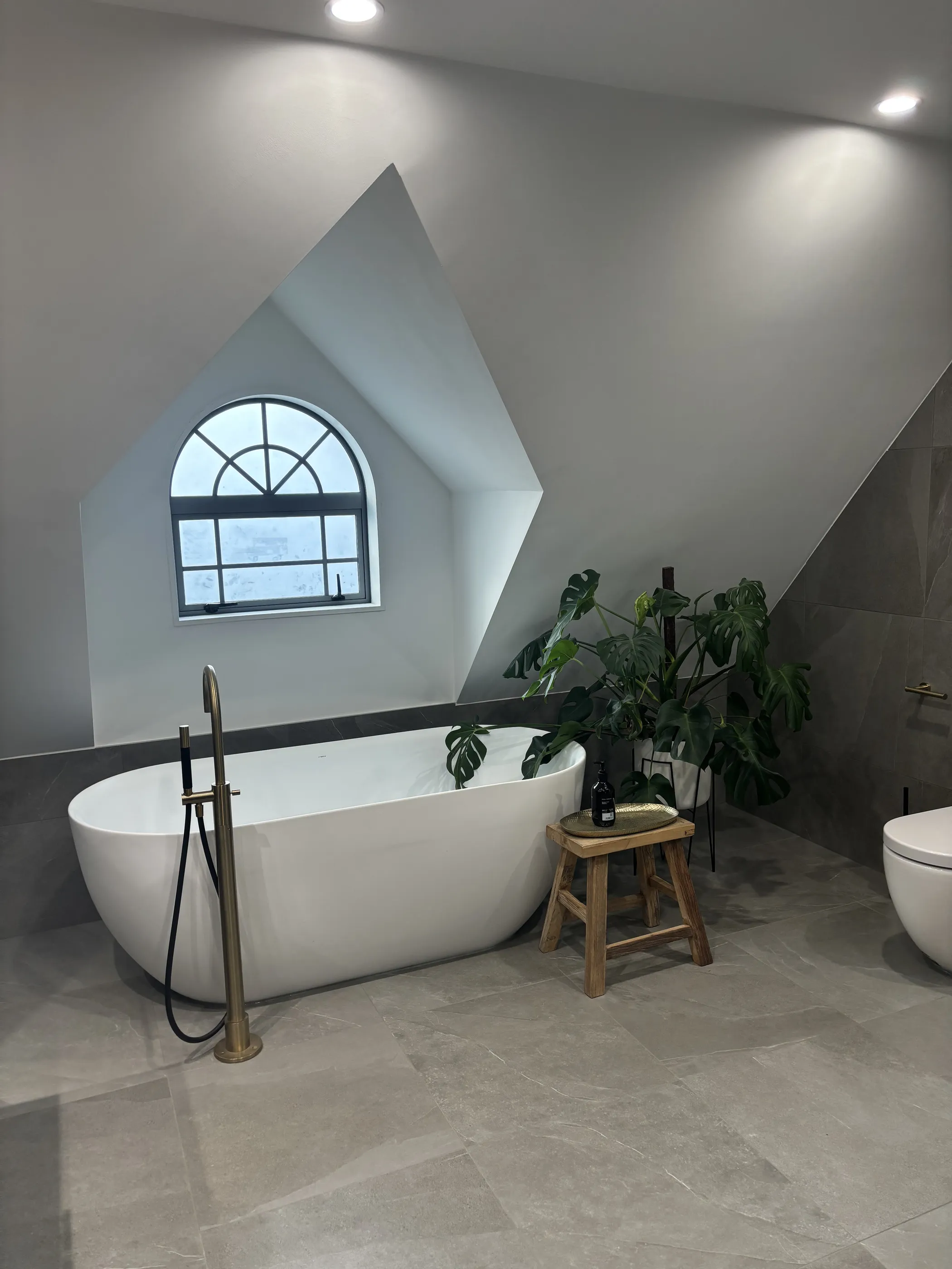 An attic bathroom with a freestanding bath under an arched multi-pane window set into a gabled reveal, with a brass floor-mounted bath filler.