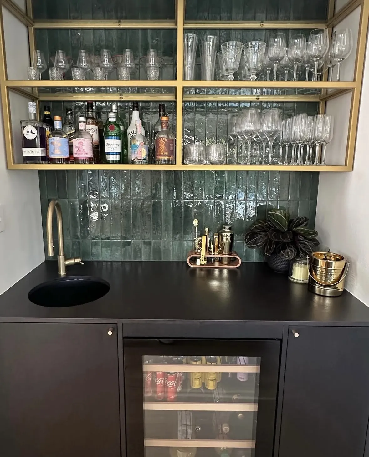 A bar with black cabinetry and a brass-framed glass shelf of glassware above, set against deep green stacked tile with brass tapware.