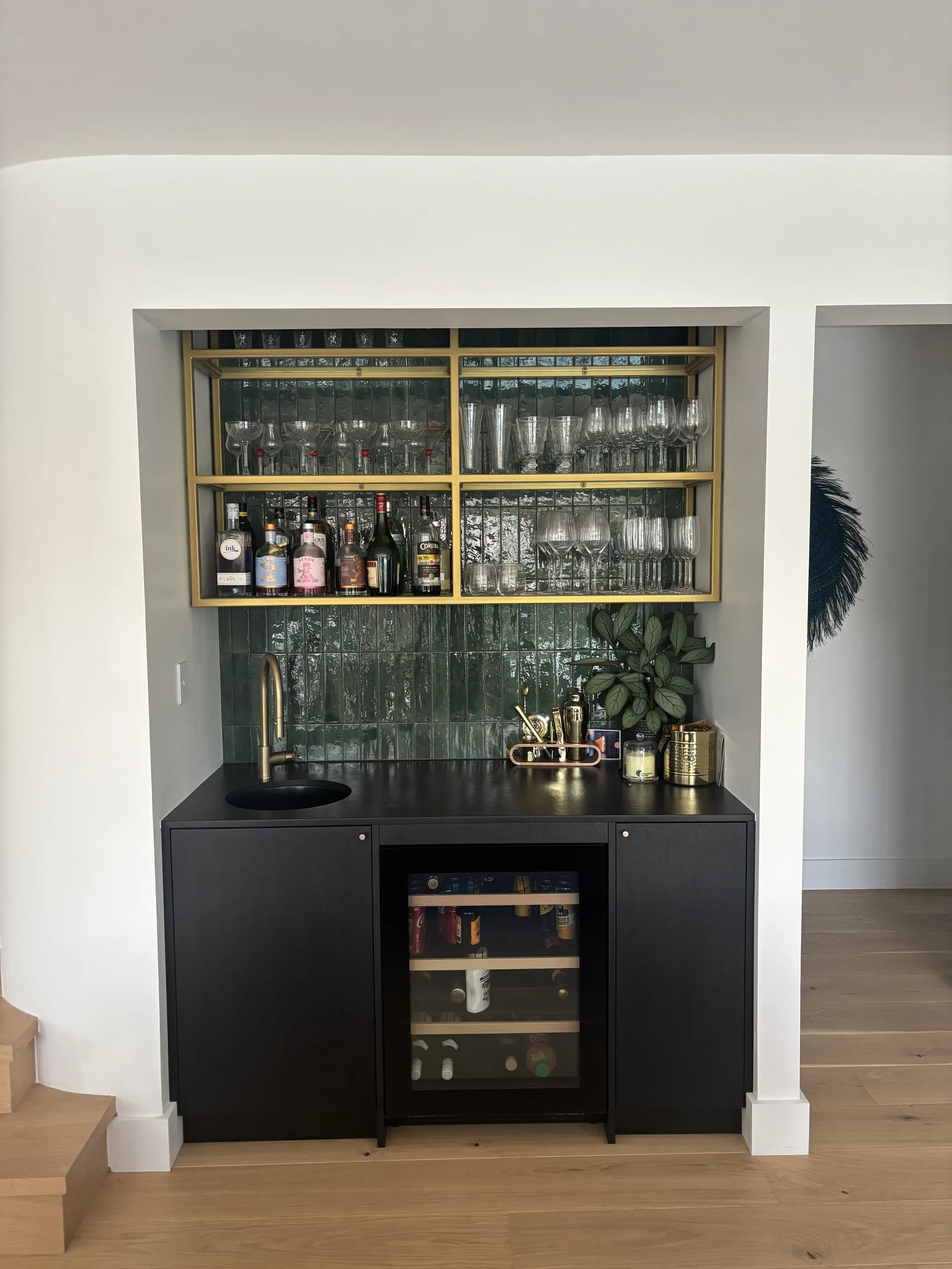 The bar seen in context, its black joinery and brass shelving set into a recess beside the stair, with oak flooring running through.