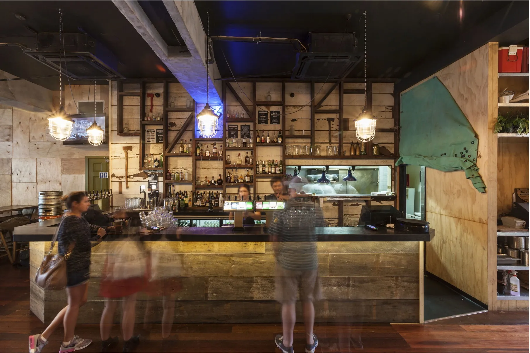 The main bar, with pendant lighting over the counter, shelves of bottles behind and plywood detailing along the walls.