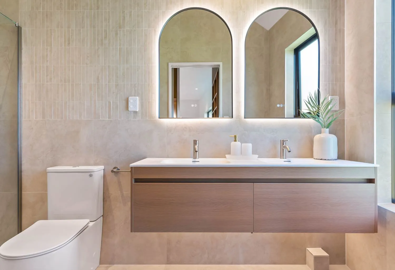 A bathroom in travertine-toned tile, with twin arched backlit mirrors above a timber vanity and a double basin.
