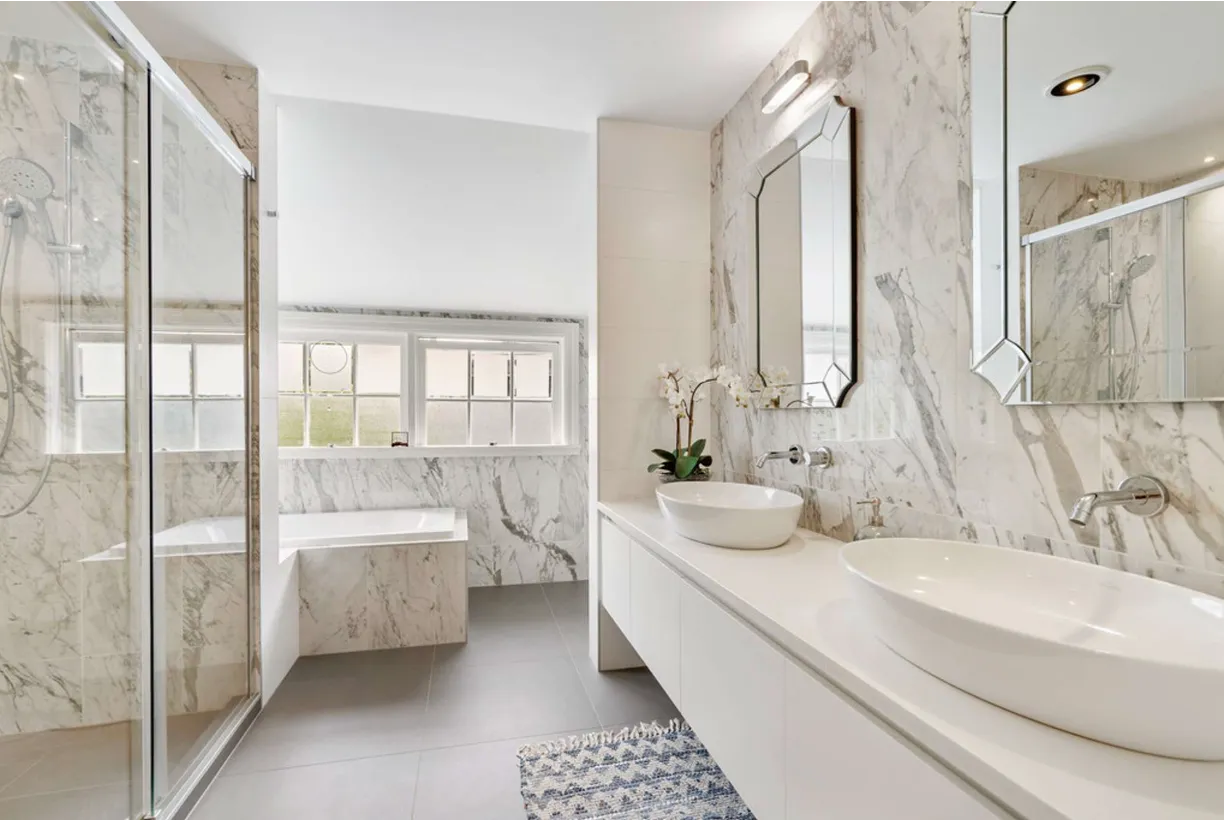 A bathroom lined in white marble, with twin vessel basins on a white vanity, faceted mirrors and a bath set into a window nook.