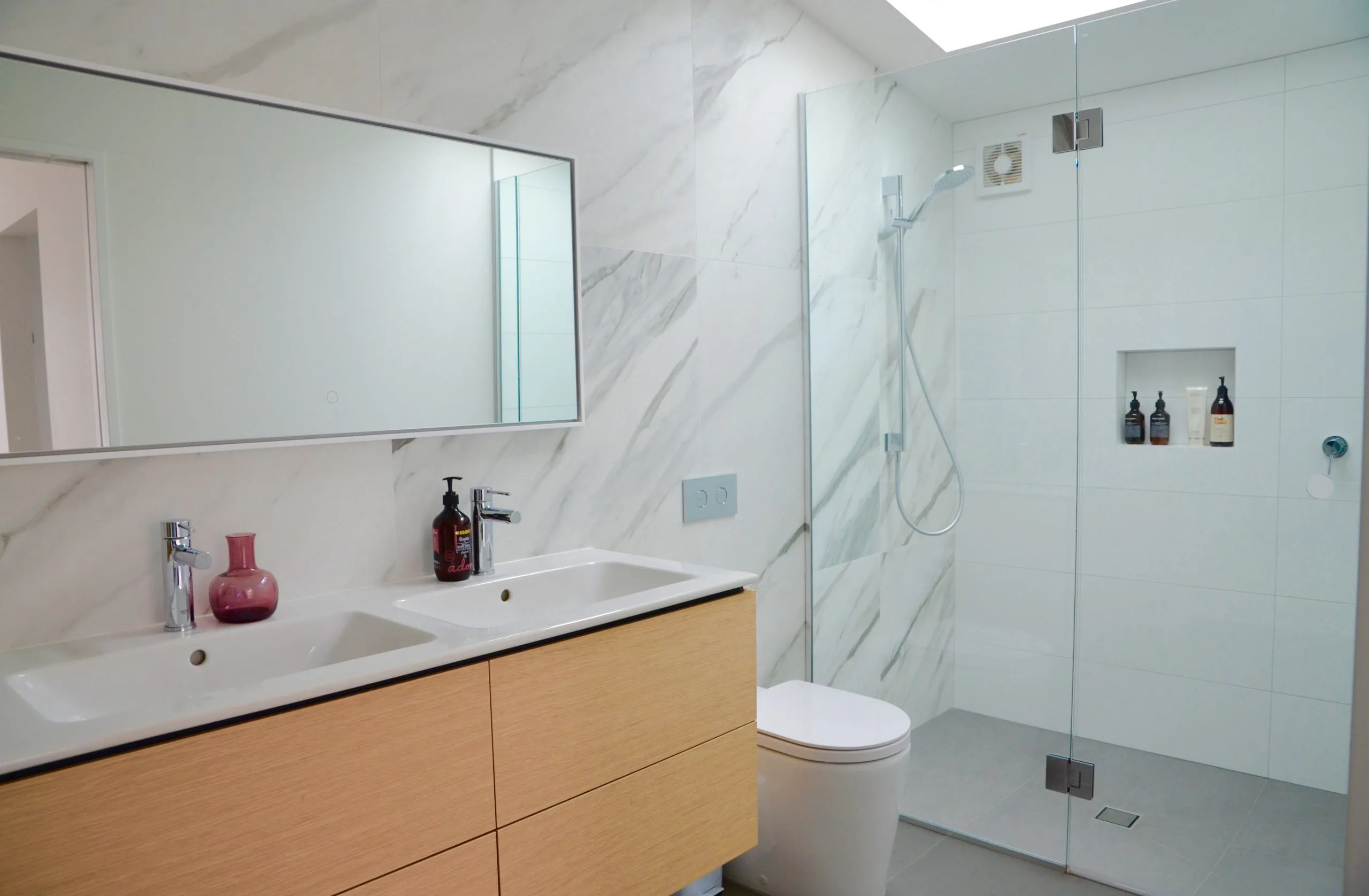 A bathroom lined in large format marble, with an oak vanity, a white basin and a round mirror.
