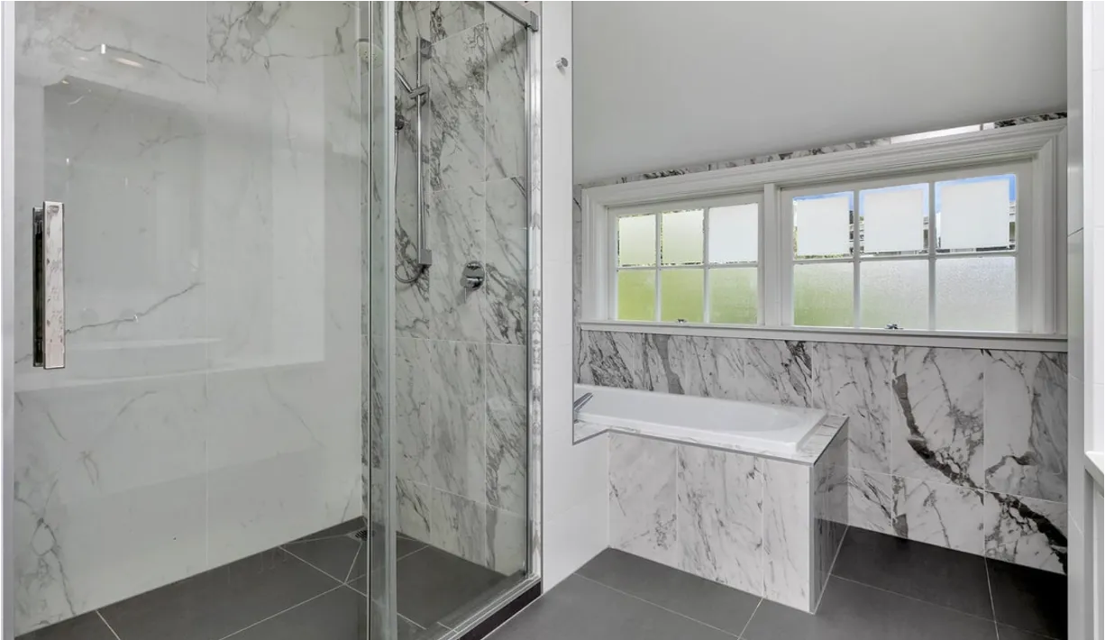 A second marble bathroom with a walk-in shower and a bath set beneath a run of multi-pane windows.