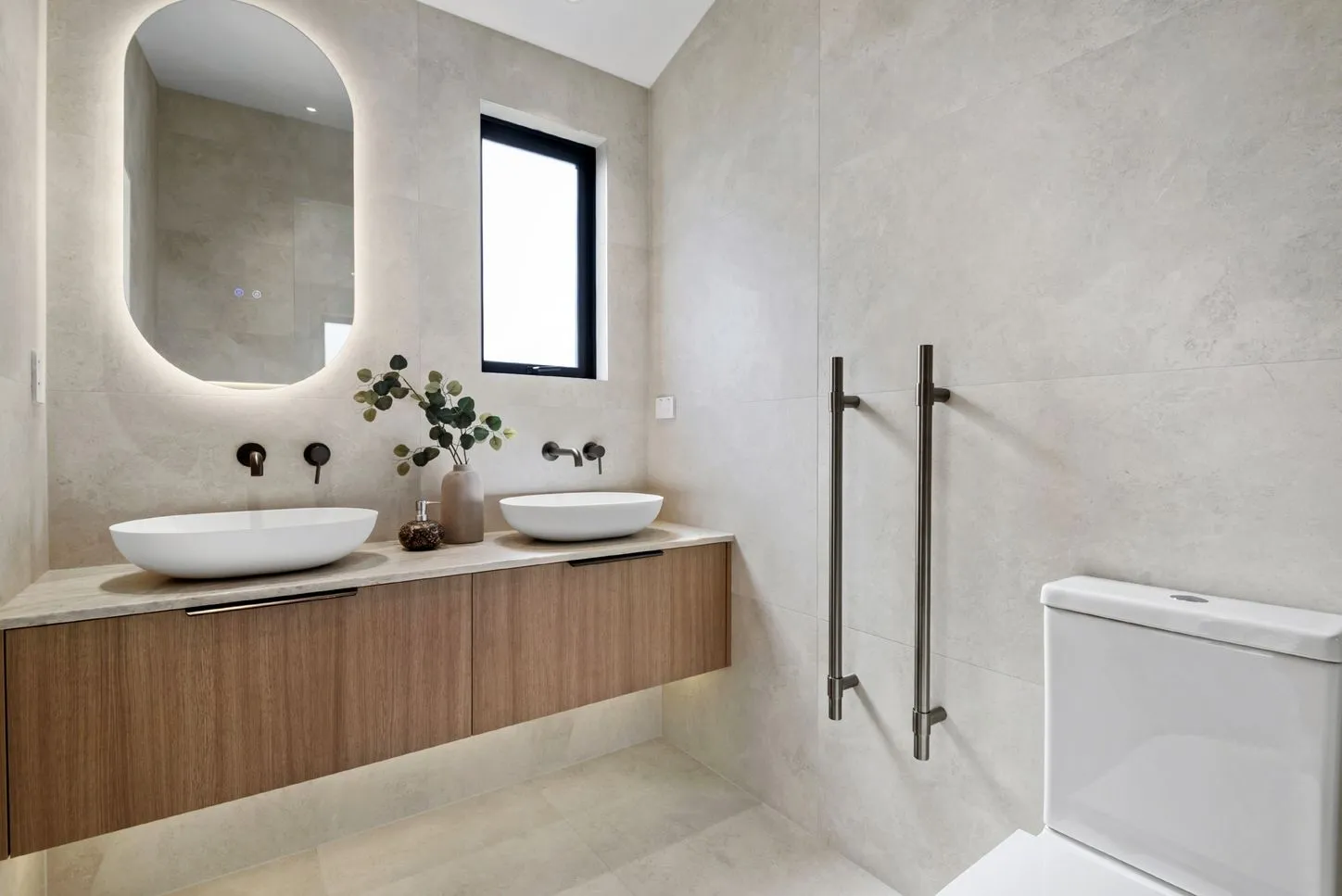 A bathroom with a backlit oval mirror, twin vessel basins on a timber vanity and large-format travertine-toned tile.
