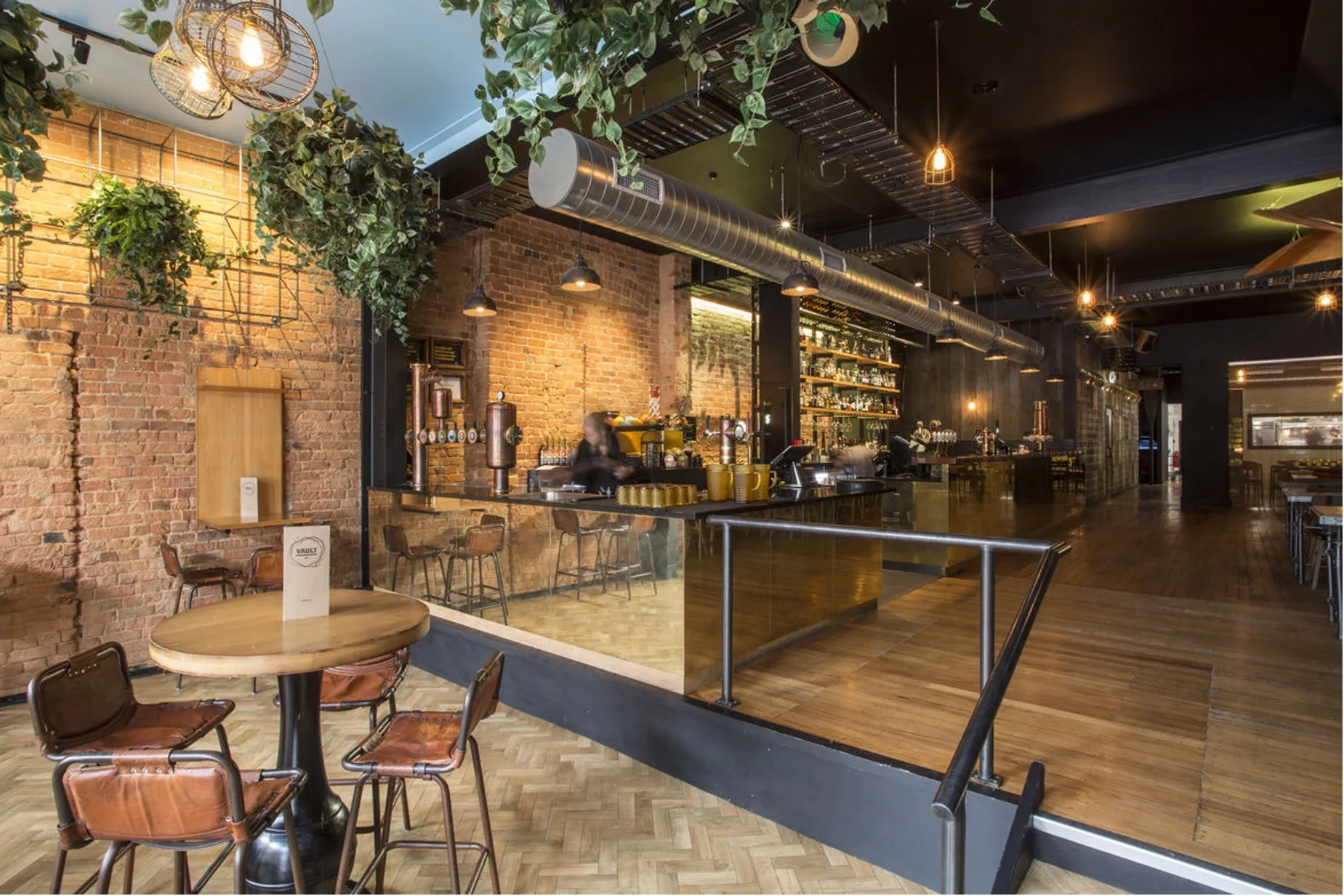 A dining area against exposed brick, with trailing greenery overhead, timber tables and leather chairs on a herringbone floor.