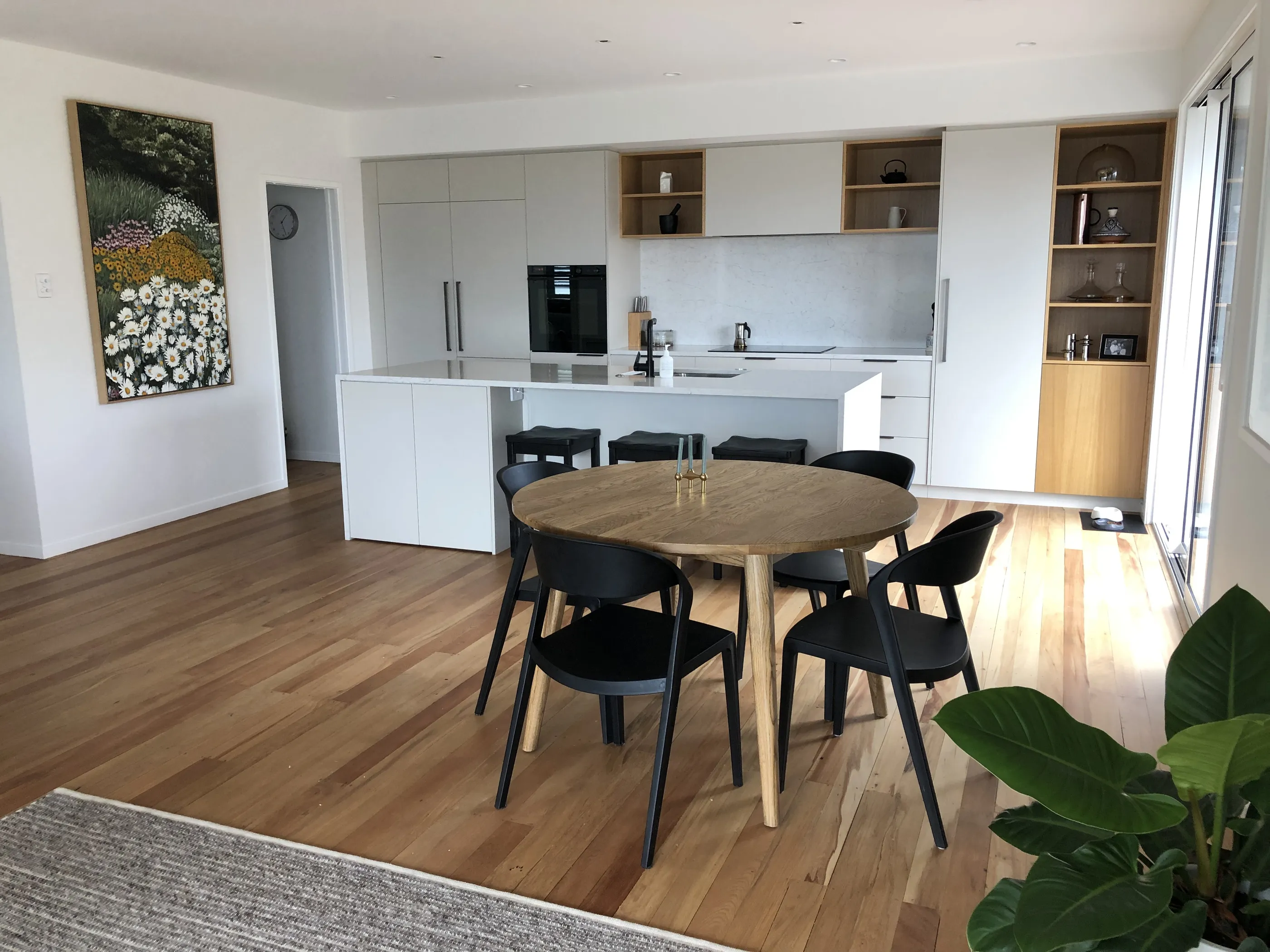 A round oak dining table with black chairs on an oak floor, the kitchen visible beyond and artwork on the wall.
