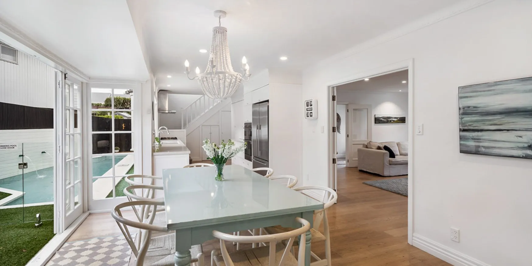 A white dining room with a beaded chandelier above a pale table and wishbone chairs, French doors folded open to the swimming pool and lawn beyond.