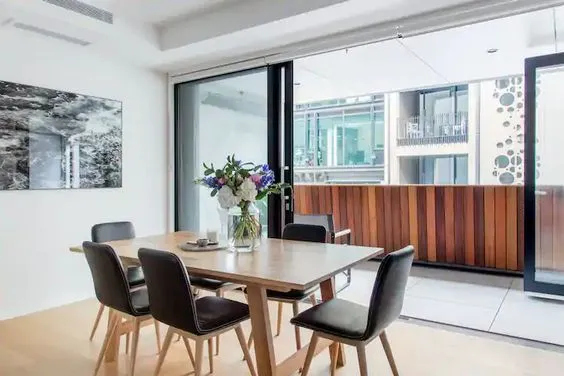 A dining area with a timber table and dark upholstered chairs, framed artwork on the wall and timber joinery running behind.