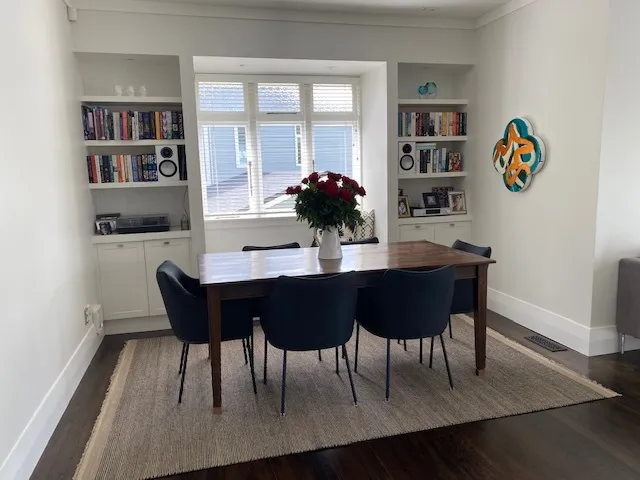 A dining room with a timber table and upholstered chairs, set between built-in bookshelves either side of the window.