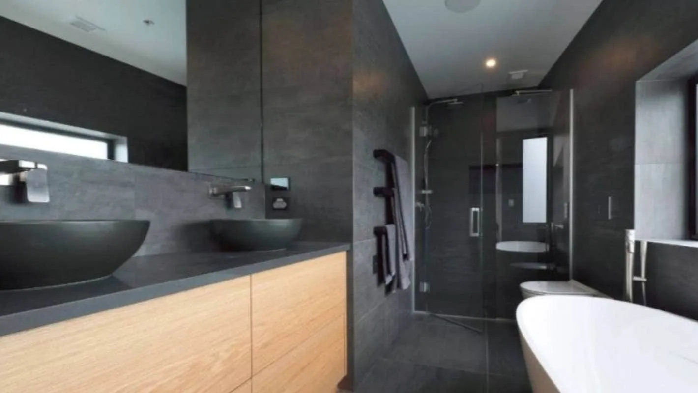 A bathroom in dark stone with twin stone basins on a timber vanity, a walk-in shower and a freestanding bath beyond.