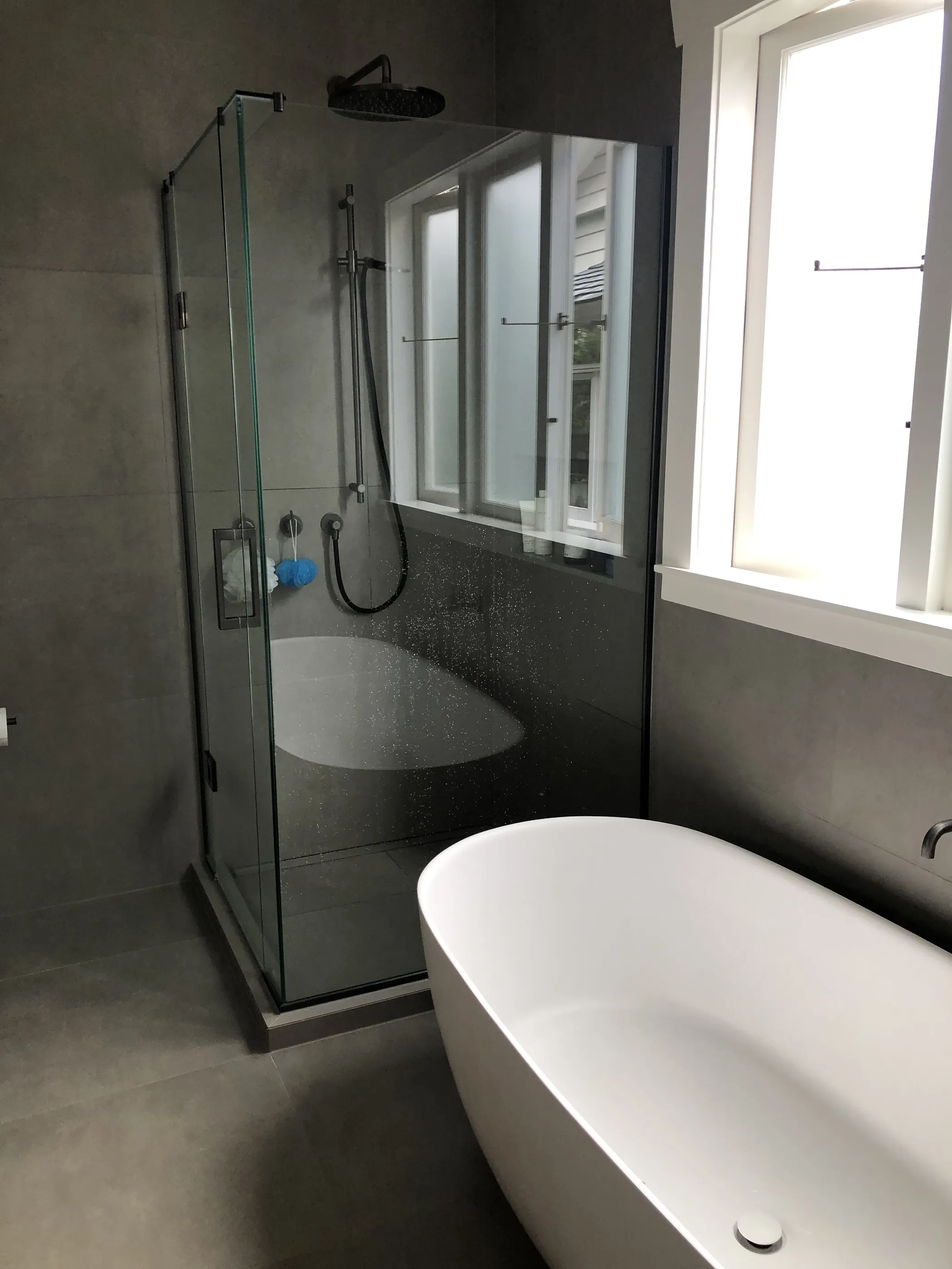 A bathroom with a freestanding bath beside a fully glazed shower, in soft grey tile.