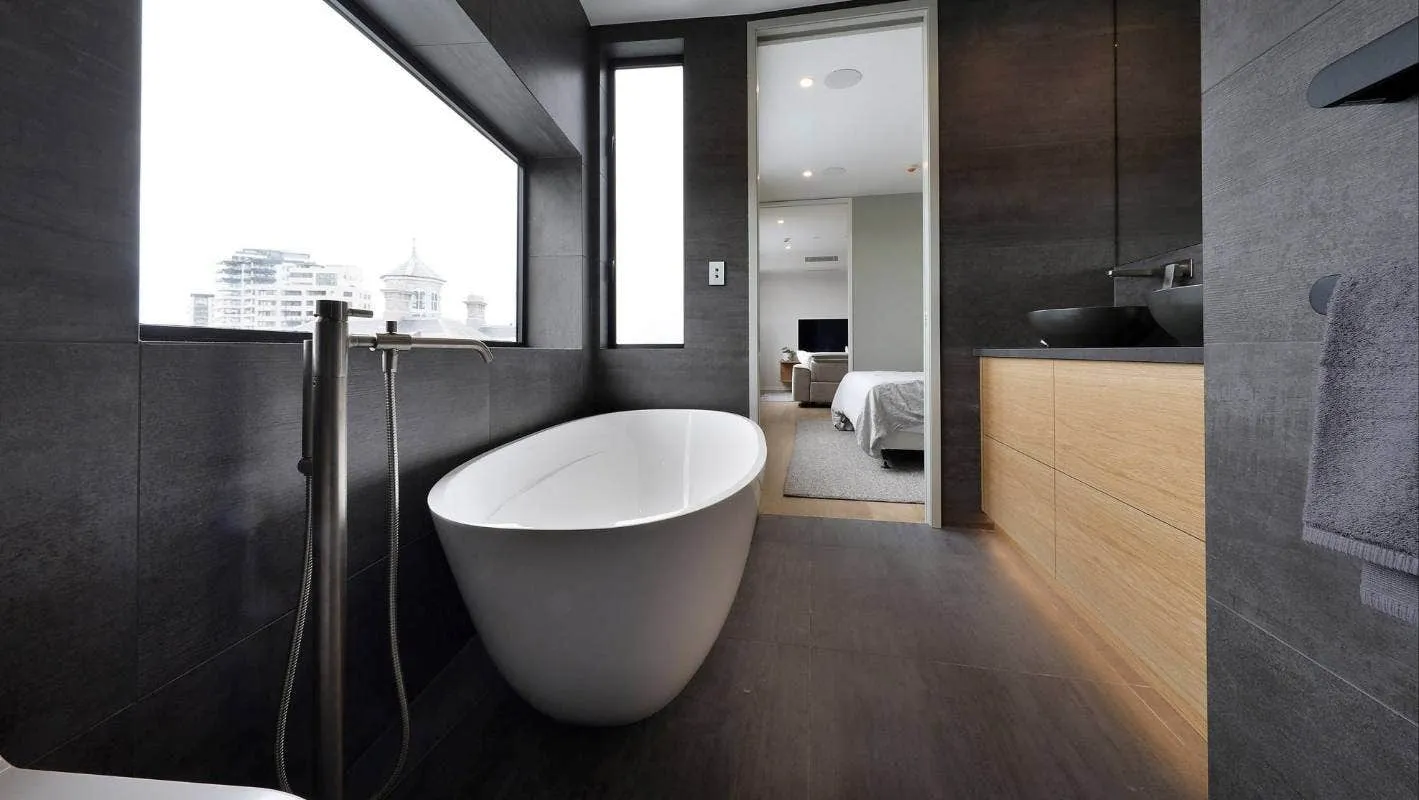 An ensuite lined in dark stone, with a freestanding bath set below a window looking out across city rooftops, and the bedroom visible beyond.