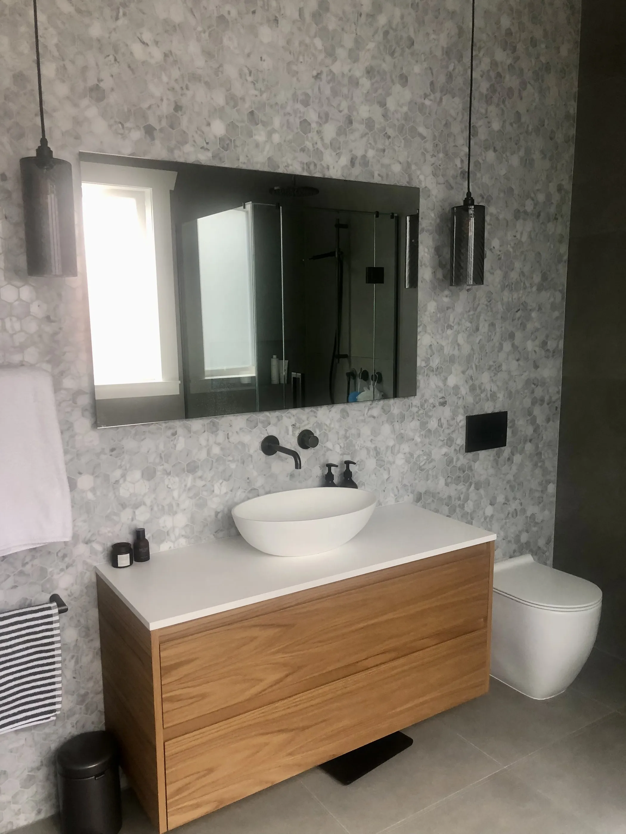An ensuite with grey mosaic walls, a timber vanity, a white basin and black tapware below a mirrored cabinet.