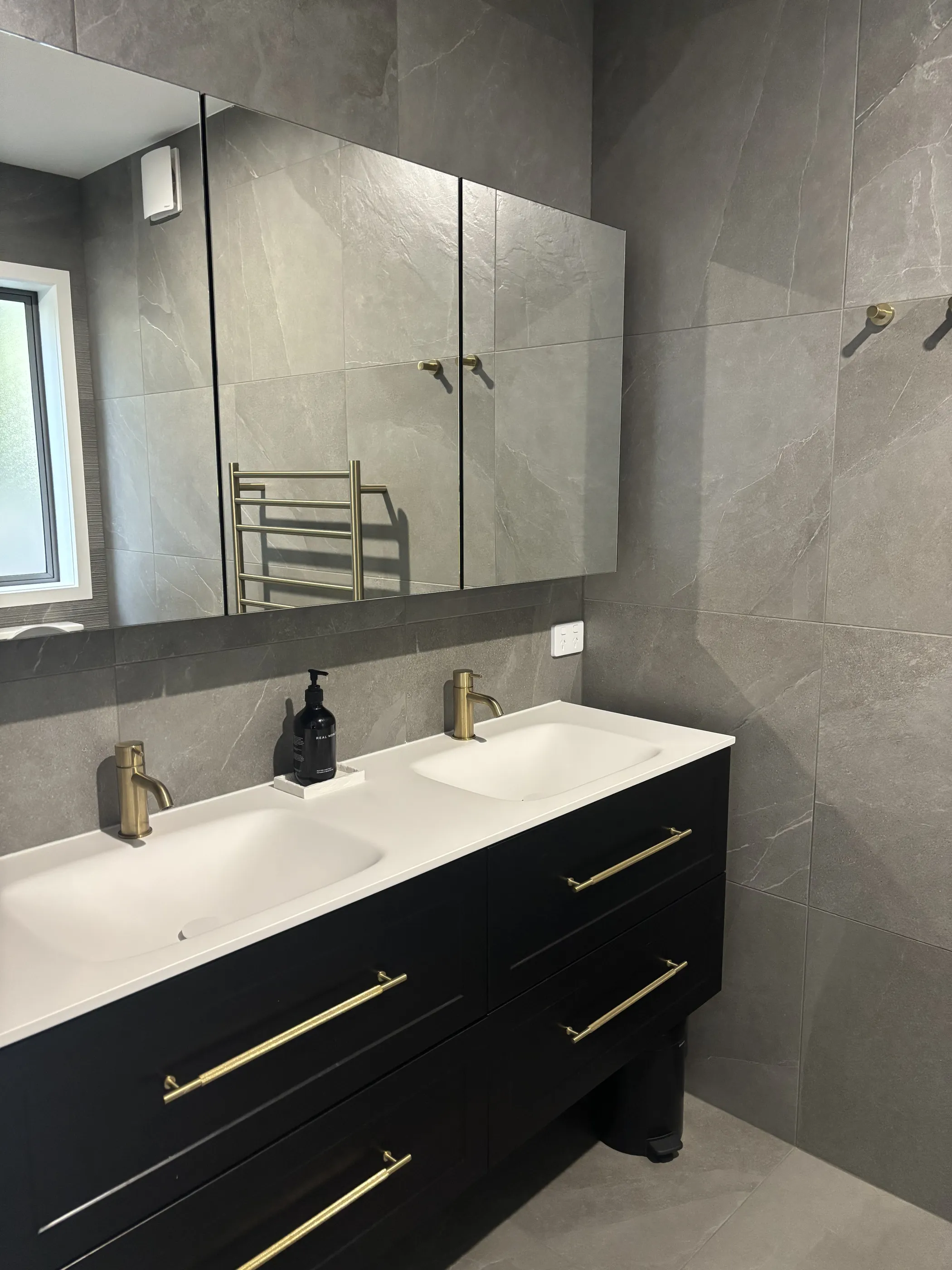 An ensuite with twin basins on a black vanity with brass handles, large format grey stone tile and a full-width mirrored cabinet.