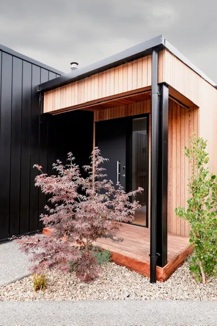 The entry, a cedar-lined portal with a black door, a hardwood step and a Japanese maple in gravel alongside.