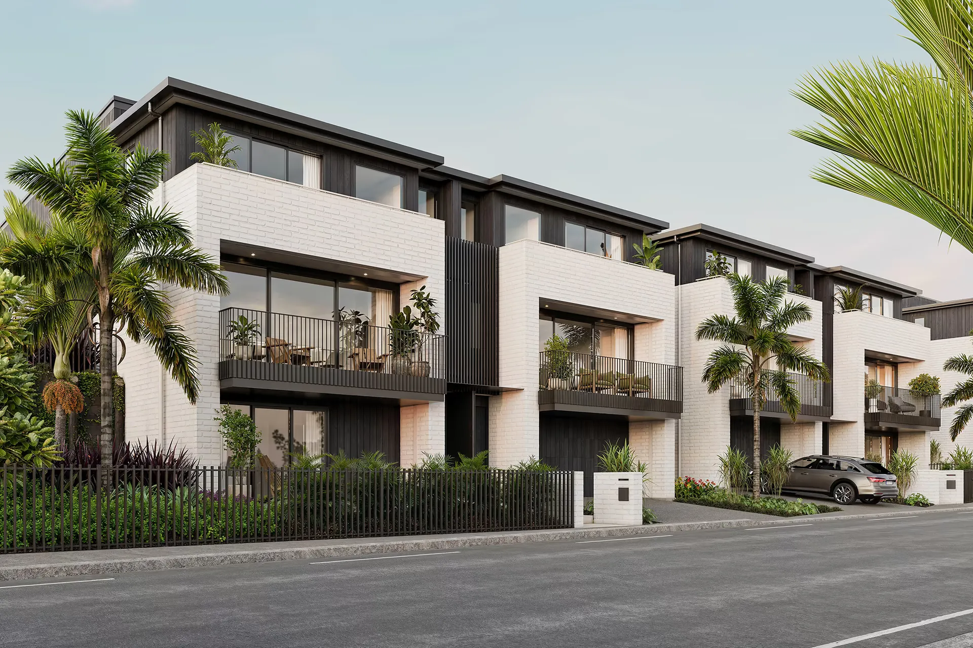 A street elevation of a terrace of duplex homes in white brick and dark vertical cladding, with balconies, palms and dense planting along the frontage.
