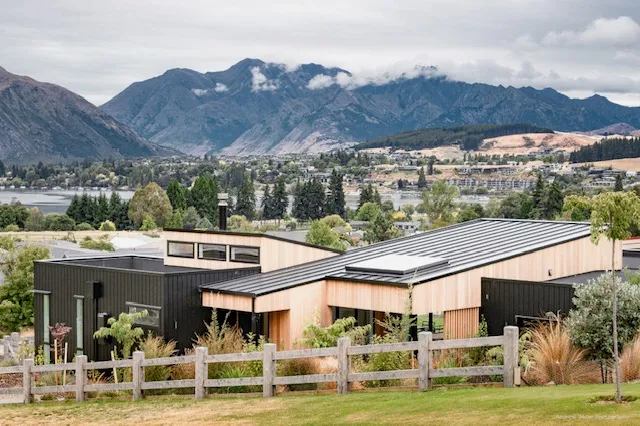 A low black-clad house with cedar detailing, set in tussock and native planting, with the lake and mountains behind it.