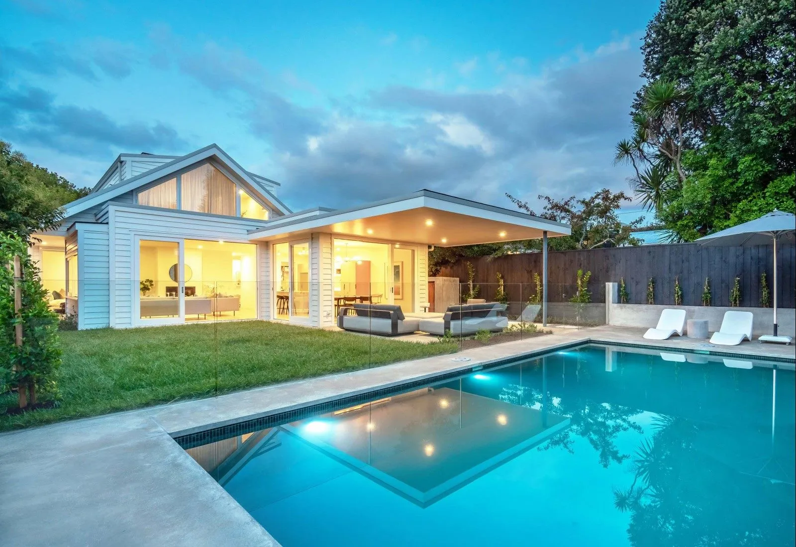 A weatherboard house at twilight with its windows lit, seen across a lap pool set into the lawn.