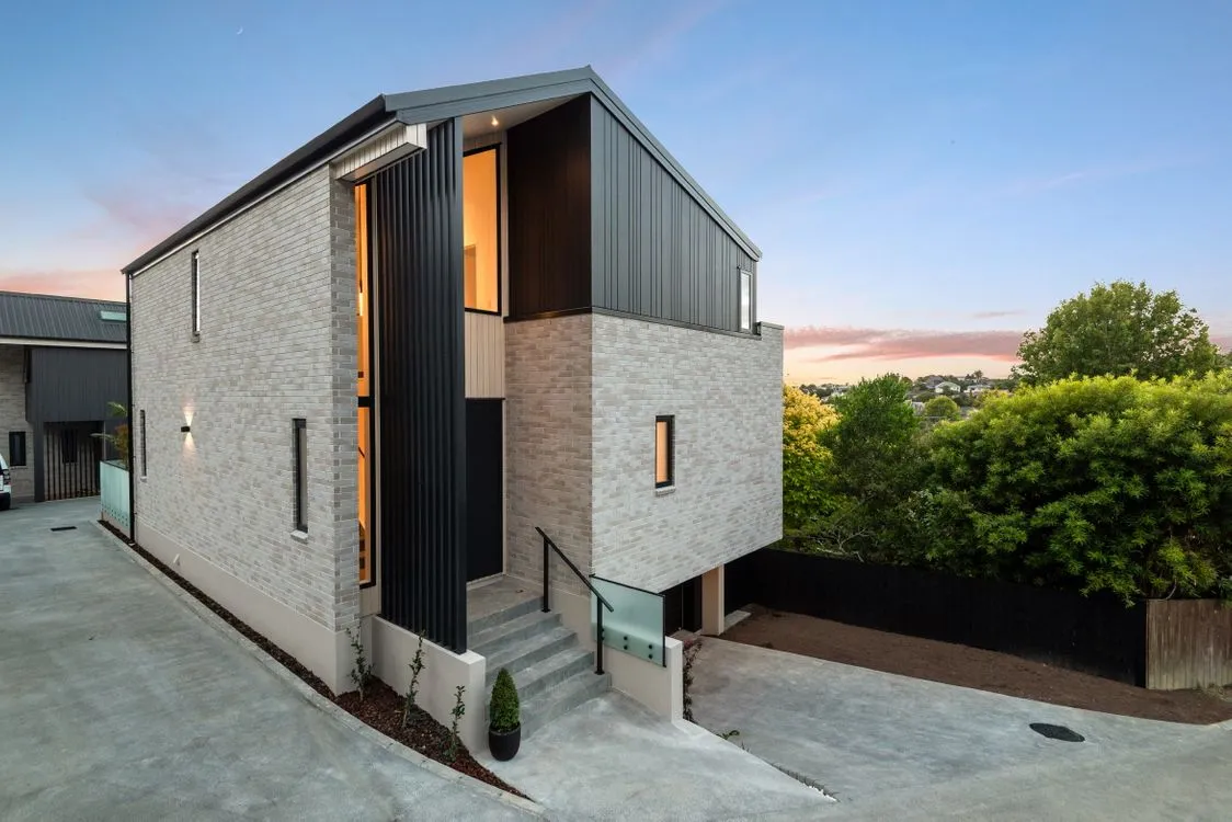 A two-storey home at twilight in pale brick and dark vertical cladding, its windows lit from within, with a glass balustrade and steps to the entry.