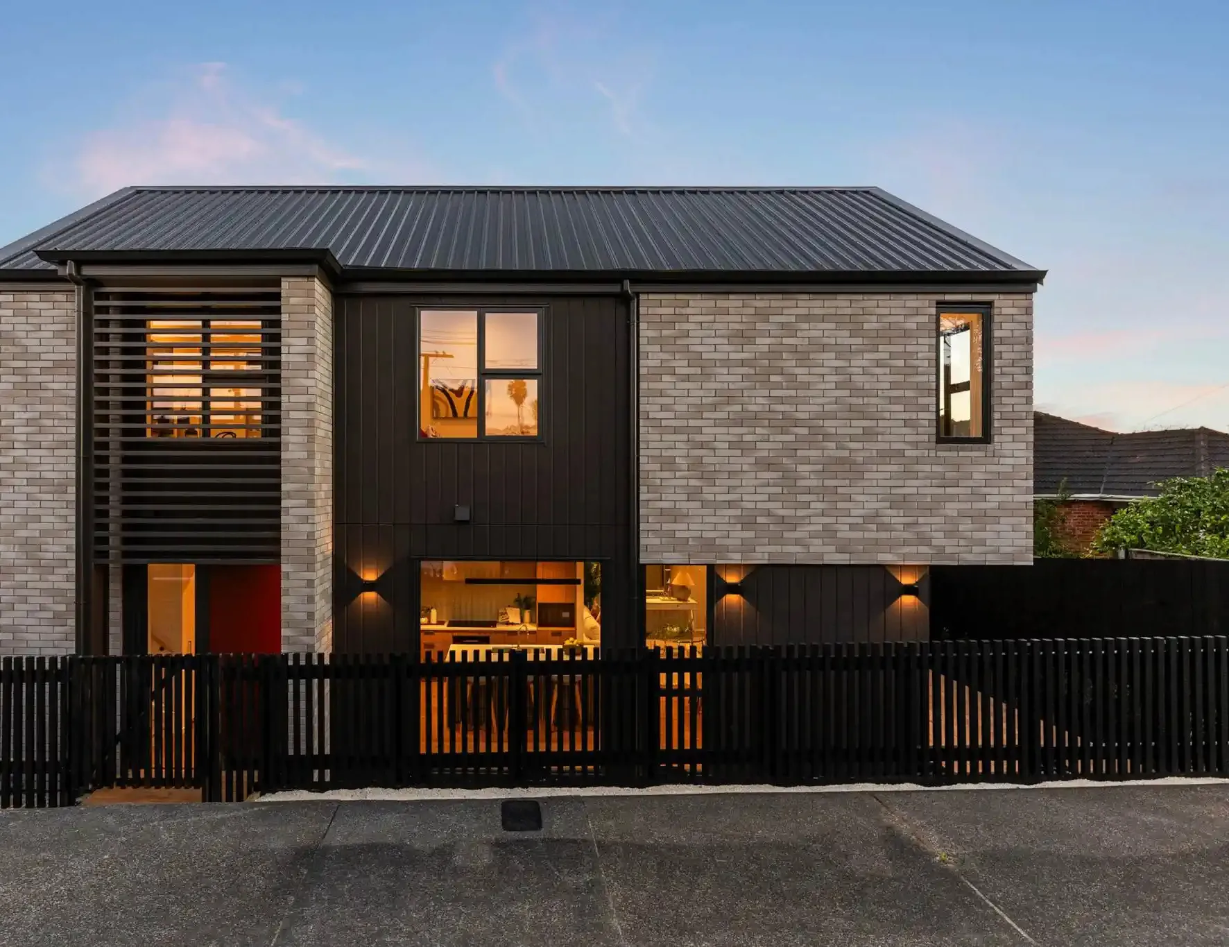 The homes at twilight, in pale brick and dark vertical cladding, with warm light showing through the ground-floor glazing behind a black picket fence.
