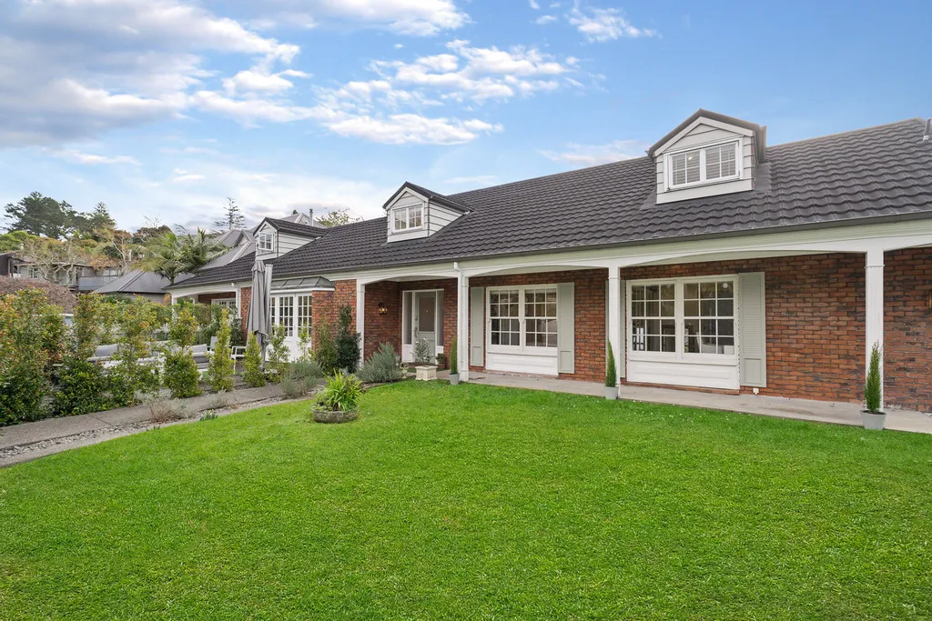 The front of a brick house with white dormer windows and a deep verandah on white posts, set behind a wide lawn.