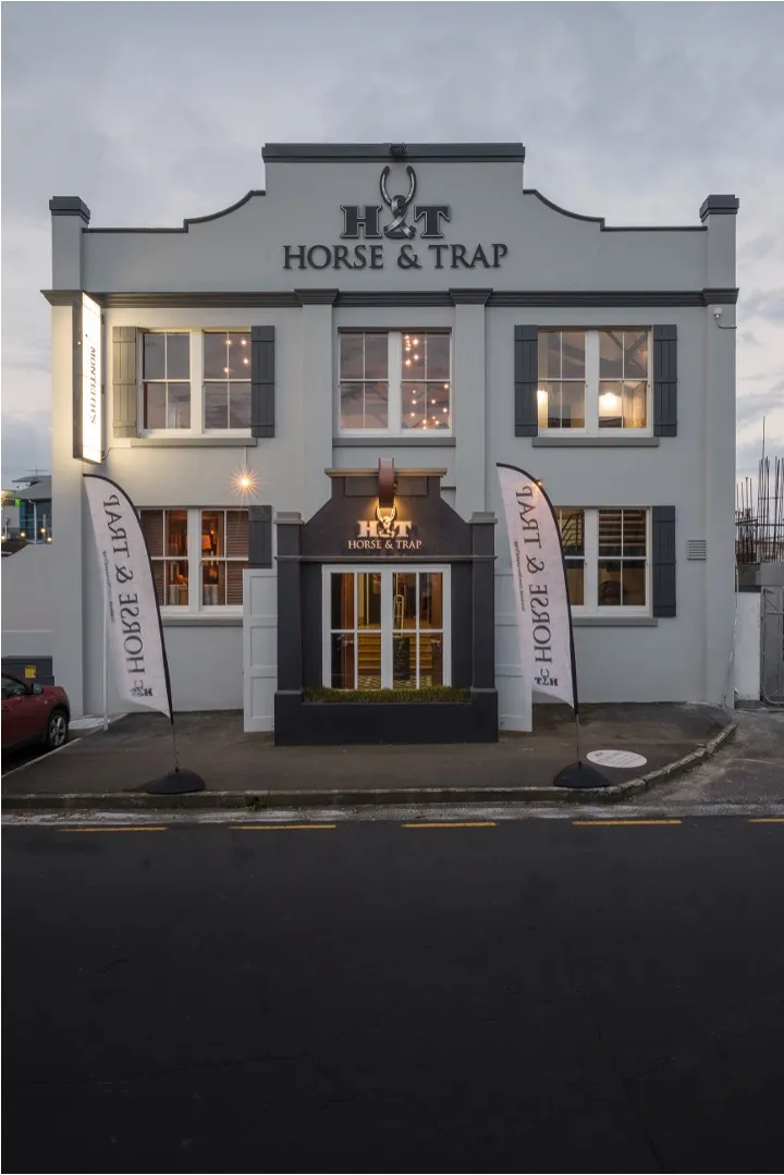 The white plastered facade of the pub at dusk, with shuttered windows lit from within and feather flags either side of the entrance.