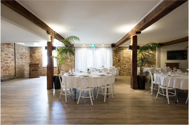A function room with exposed brick walls, timber posts and beams, set for dining with round tables in white linen.