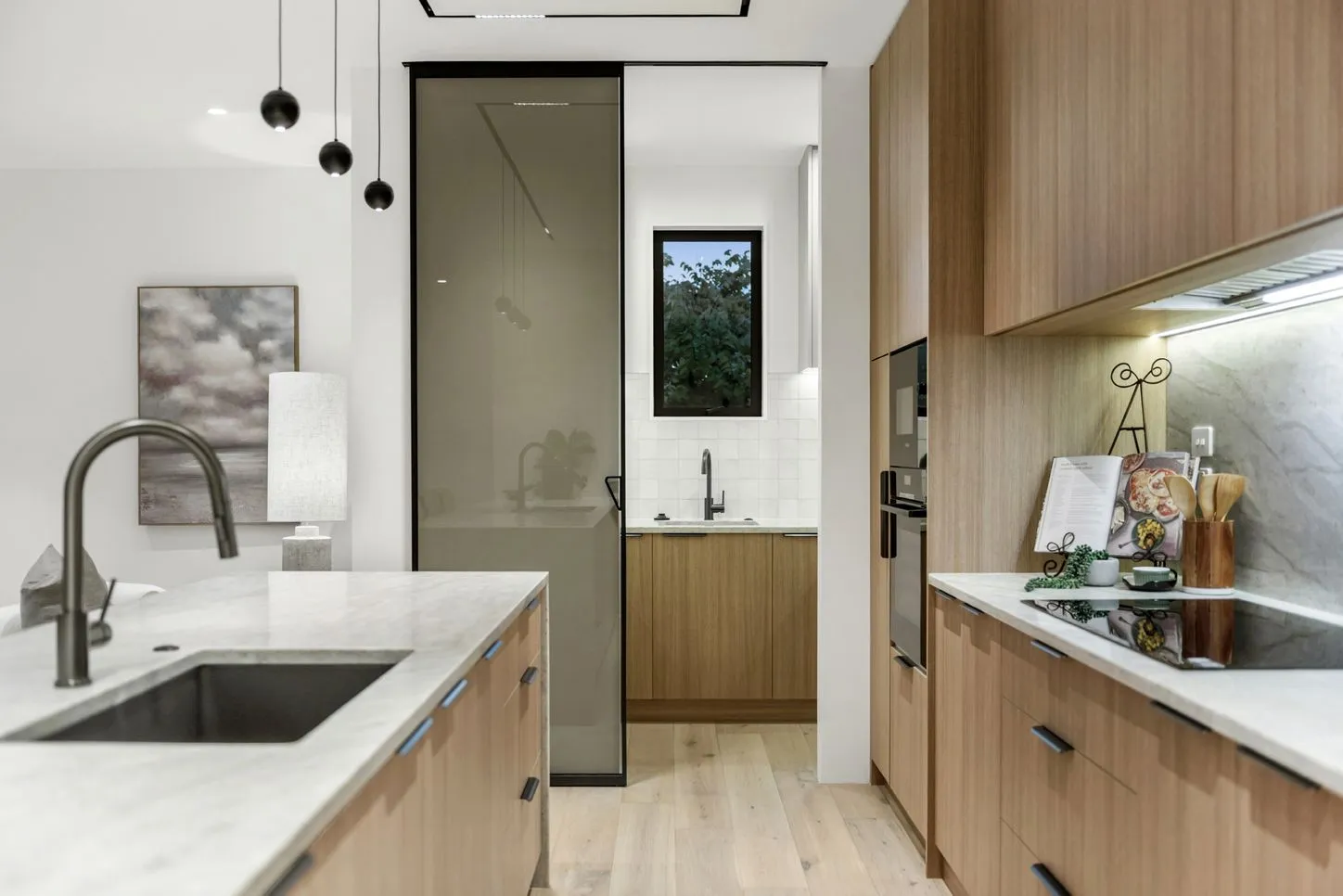 A galley kitchen and scullery in white and timber, with a window above the sink and marble splashback behind the cooktop.