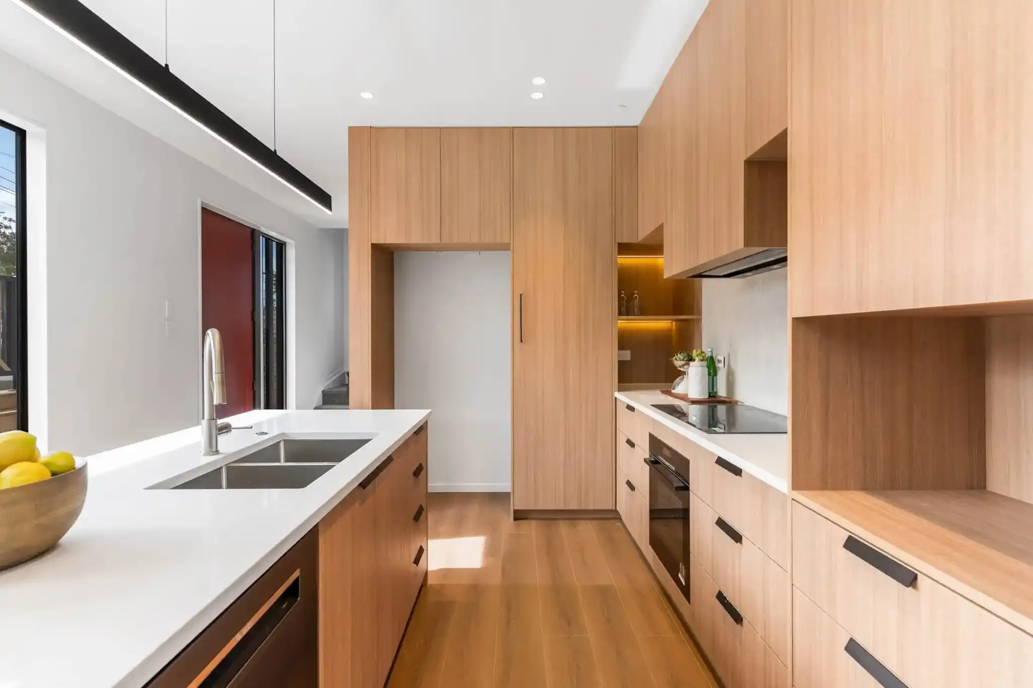 A galley kitchen with timber cabinetry and a white stone benchtop, sunlight falling across the oak floor from a high window.
