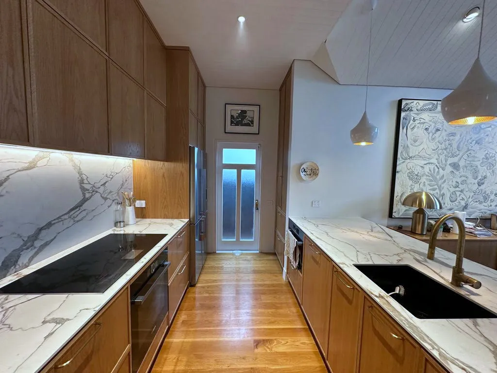 The galley run of the kitchen, with oak cabinetry, a marble benchtop and full-height marble splashback, and a brass tap over a black sink.