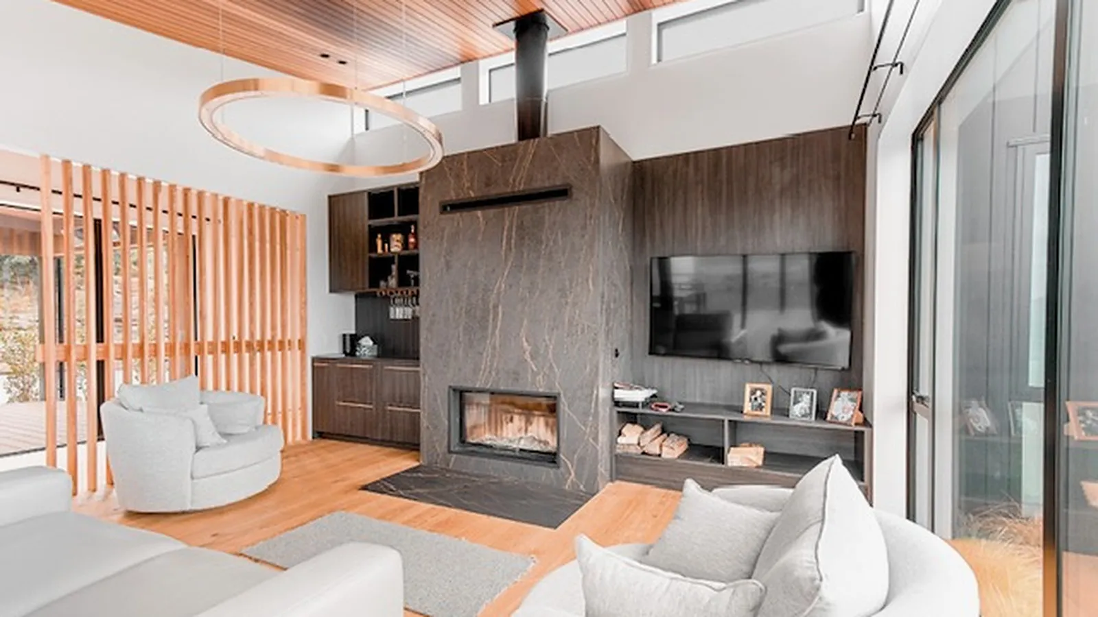 A living room under a cedar-lined raked ceiling, with a circular timber pendant, a dark veined stone fireplace, a timber screen wall to one side and pale oak flooring throughout.