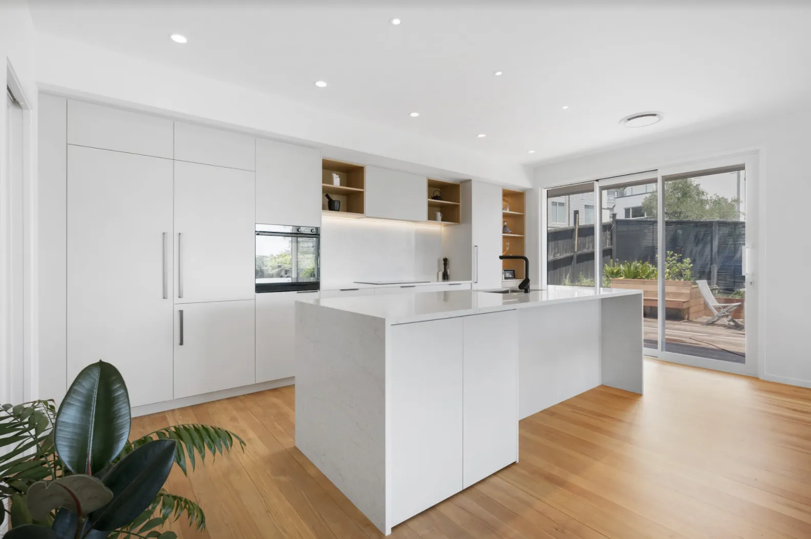 The kitchen seen wide, with a long stone island, tall white cabinetry and glazing to the garden beyond.
