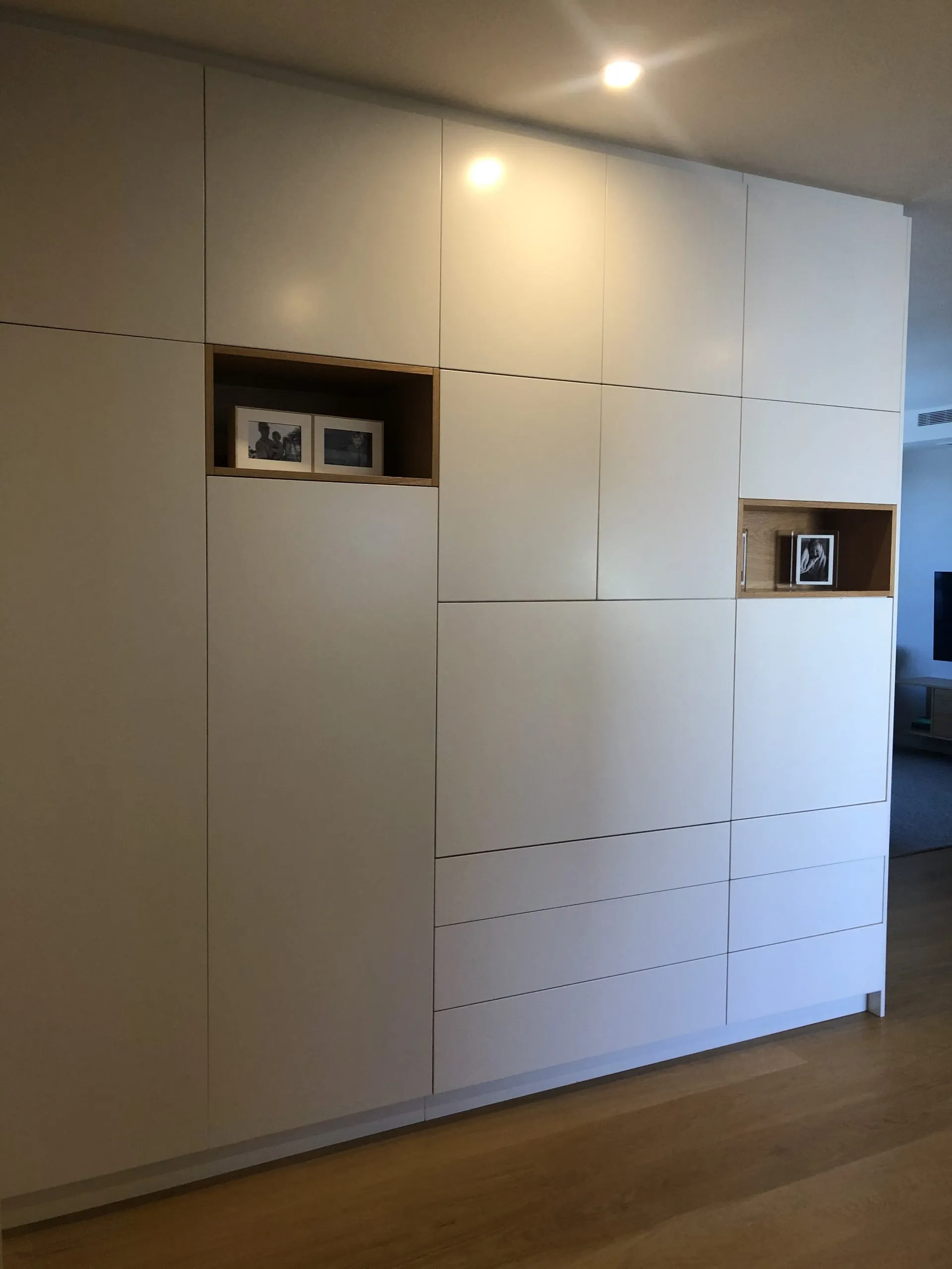 A full-height white joinery wall with oak-lined display niches set into it, running the length of the living space.