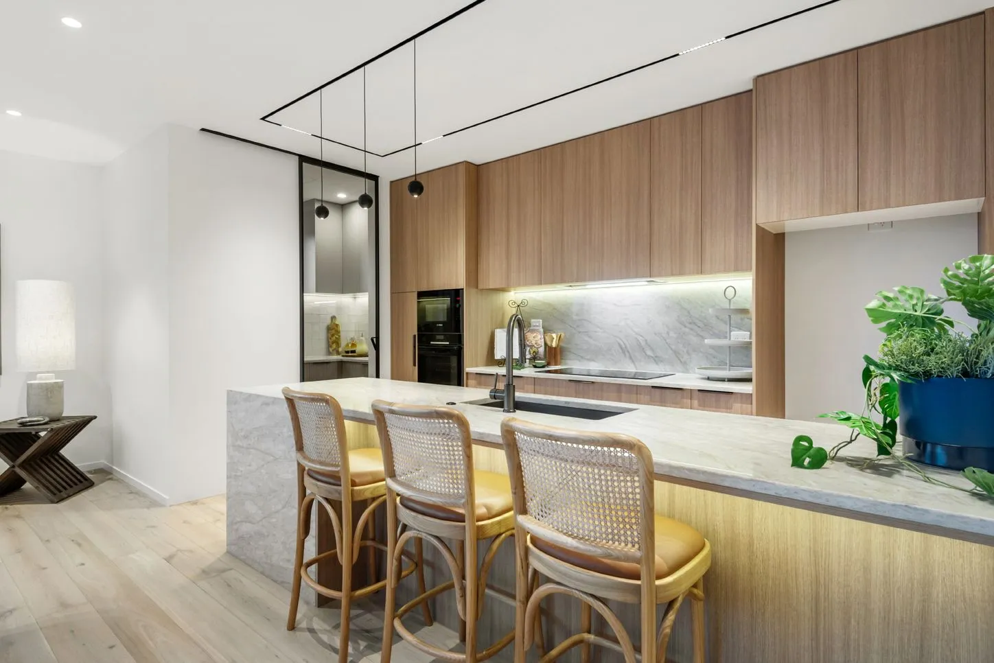 A kitchen island with cane-backed stools, a stone benchtop and planting on the counter, with living space beyond.