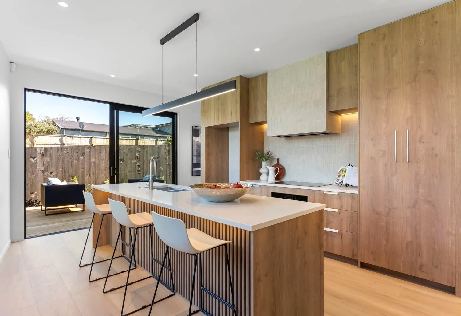 A kitchen in a different home, with timber cabinetry, a stone island and stools, opening through sliding glazing to a deck.