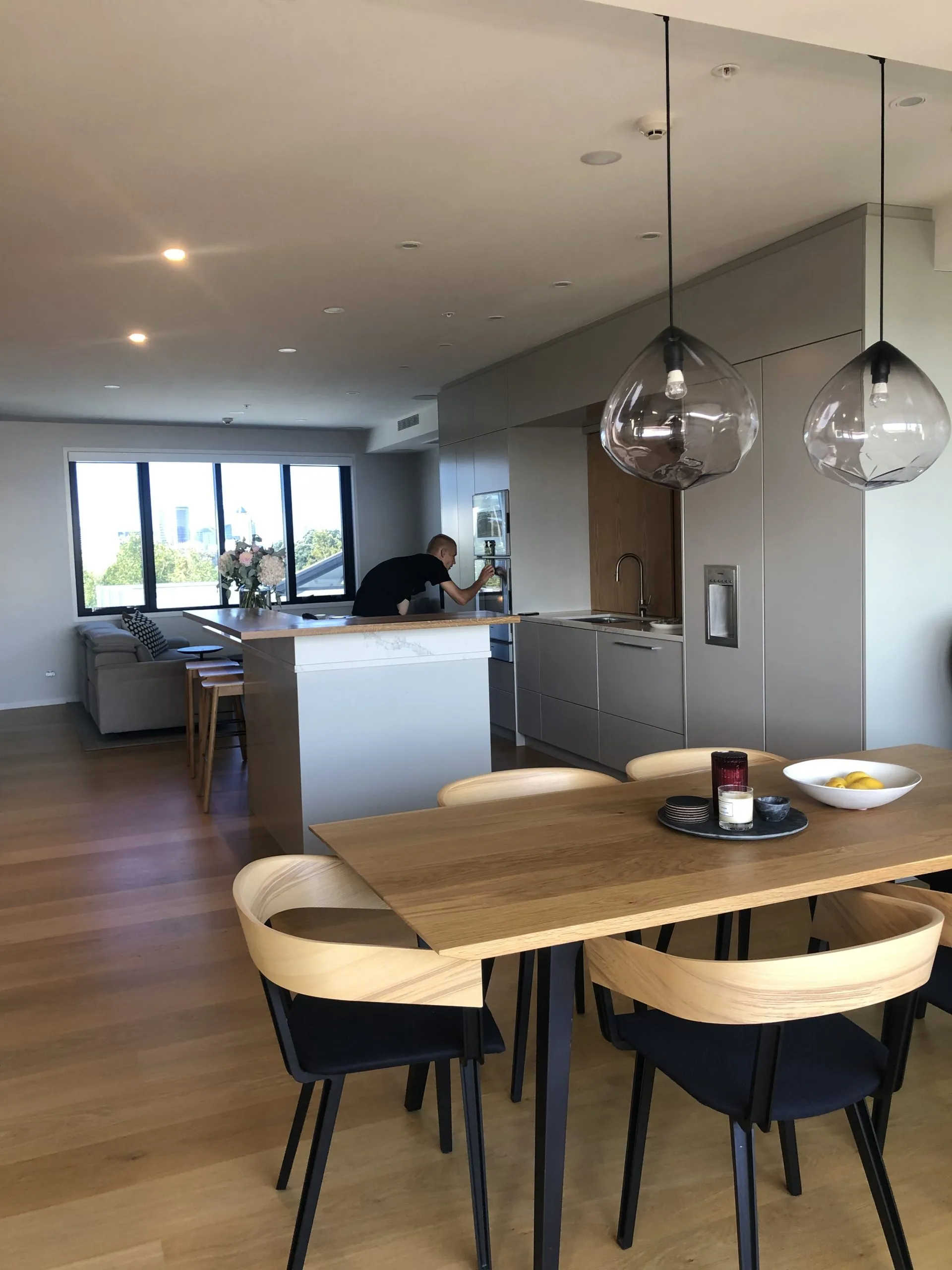 Kitchen and dining, with sculptural smoked-glass pendants above a timber table and the city visible through full-height glazing.