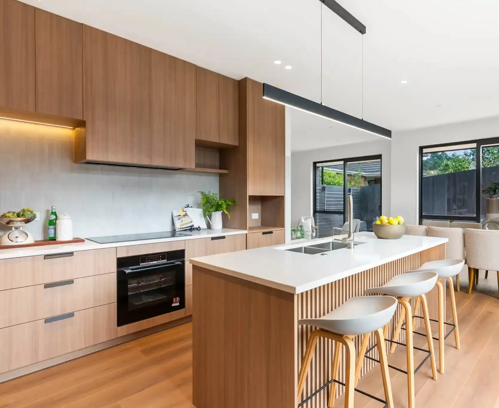 A kitchen and dining space with oak cabinetry, a white stone island on a fluted base, three stools alongside and glazing to the garden beyond.