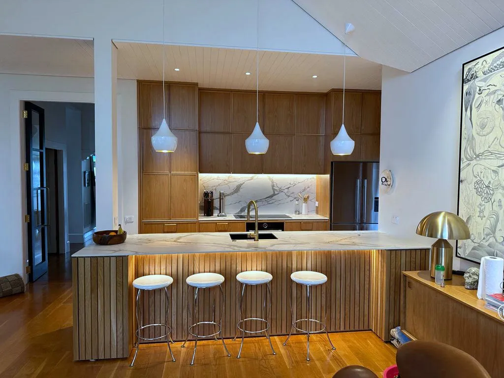 A custom oak kitchen seen wide, with a marble peninsula on a fluted timber base, stools alongside and a raked, tongue-and-groove ceiling above.