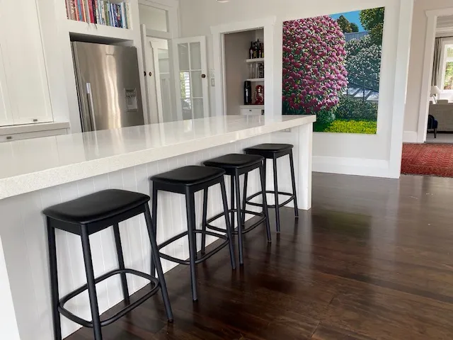 A long white stone island with black bar stools along one side, and dark timber flooring running through.