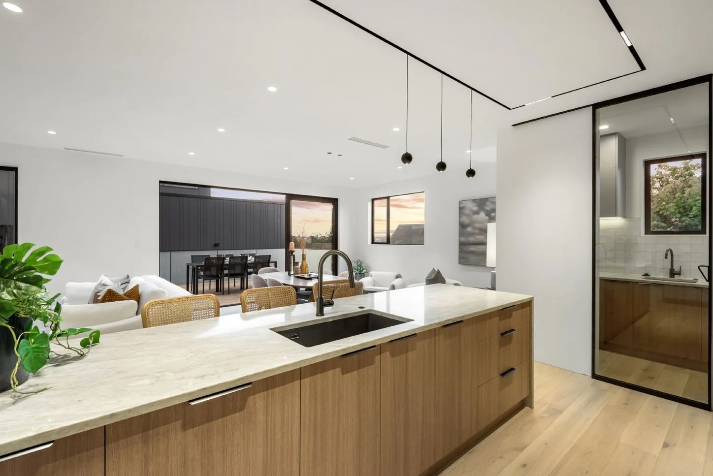 A wide view across the kitchen island to the living and dining area, with glazing to a deck and outlook beyond.