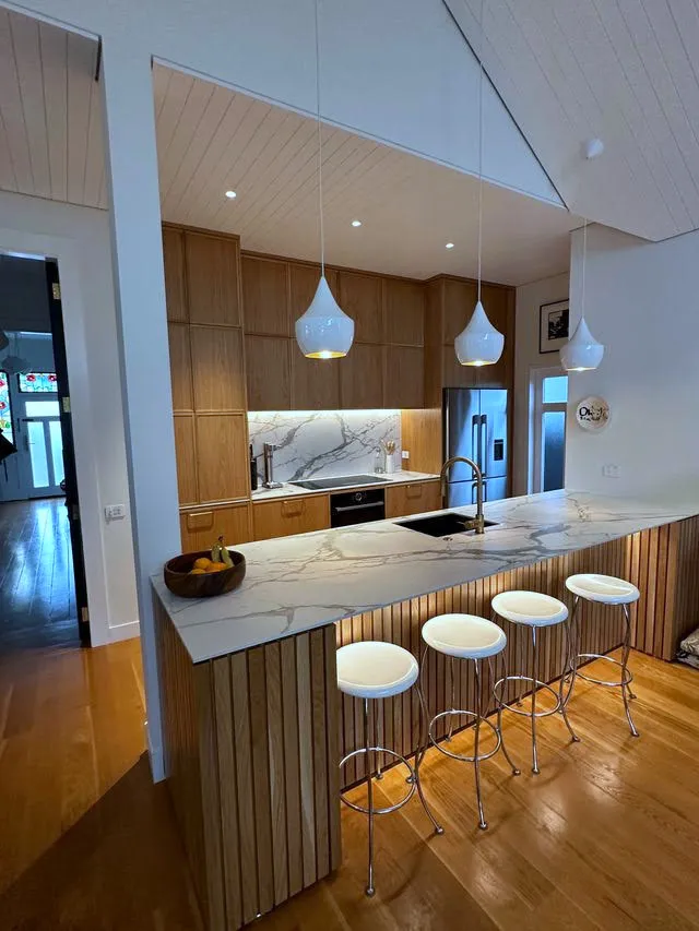 A custom oak kitchen with a marble peninsula on a fluted timber base, three white pendants above it and a raked, tongue-and-groove ceiling overhead.