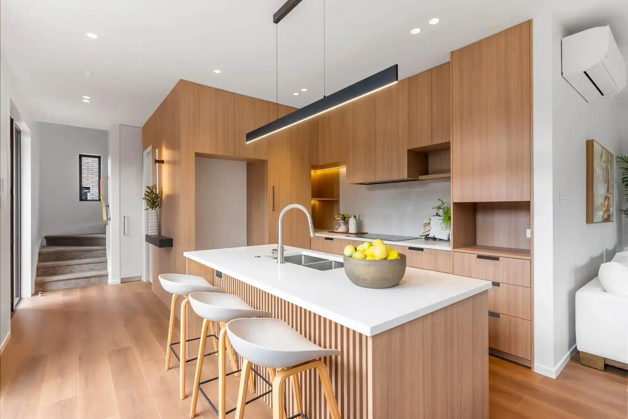 A wide view of the kitchen, with the island and stools in the foreground and the staircase and entry visible beyond.