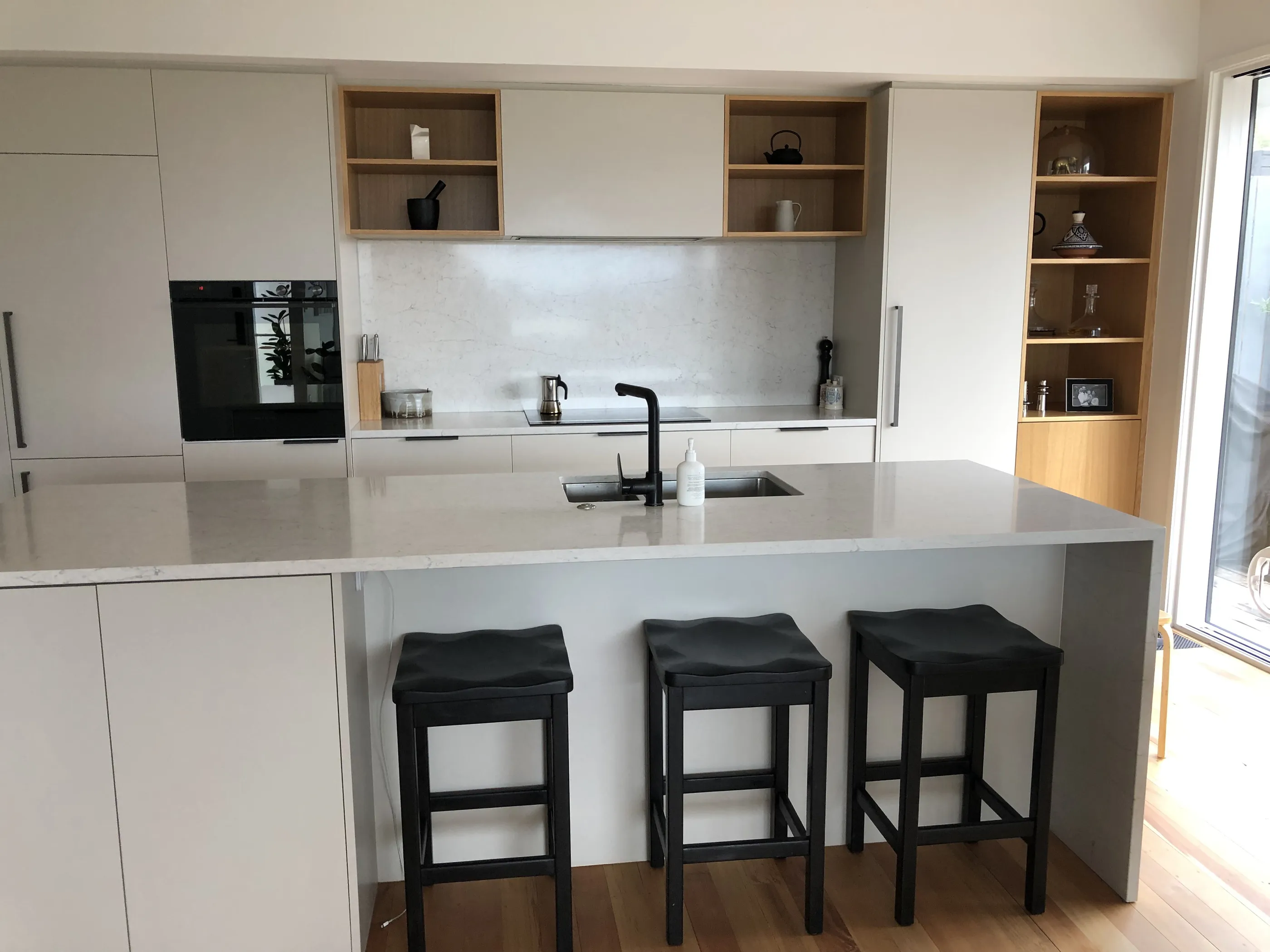 A white handleless kitchen with oak open shelving set into the run, a stone island and black leather-topped bar stools on an oak floor.