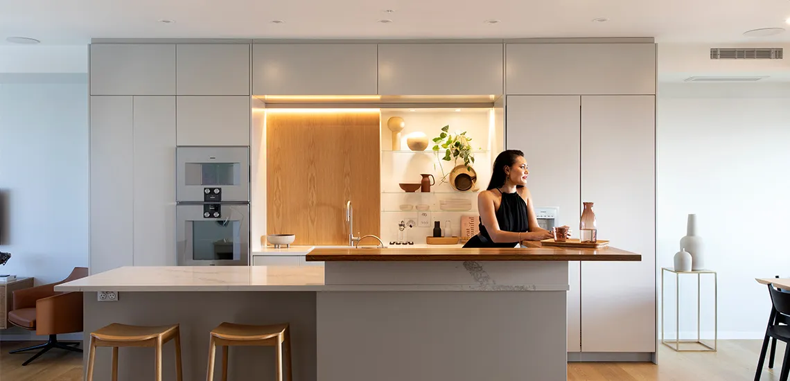 A pale kitchen with an island bench and timber counter, softly lit, with someone standing at the far end preparing food.