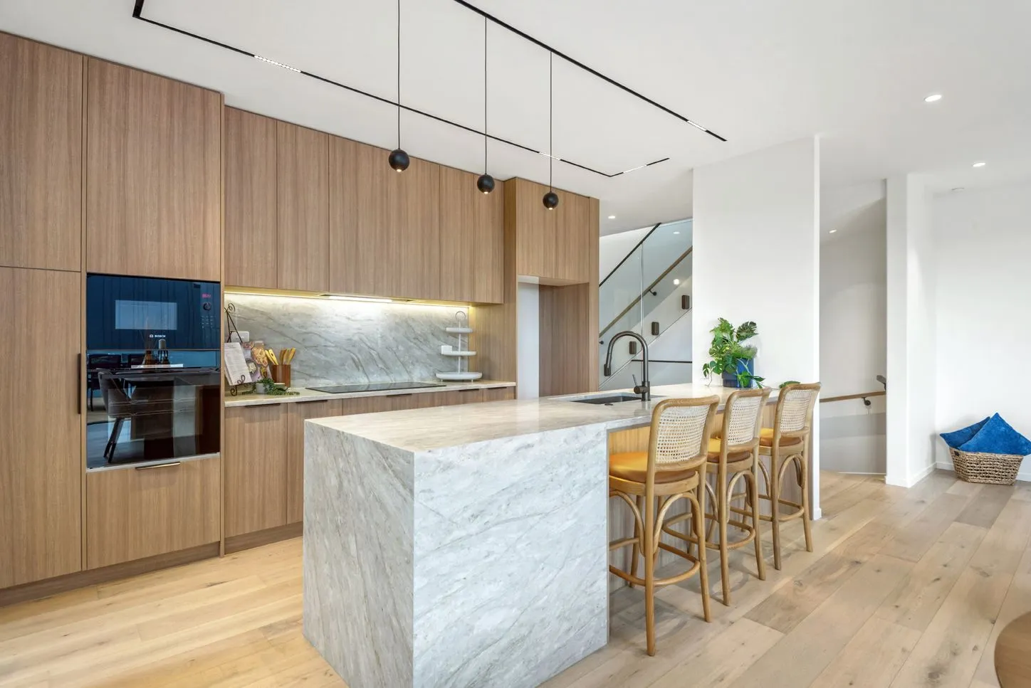 A kitchen with a marble waterfall island, oak joinery, cane-backed stools and the staircase visible beyond.