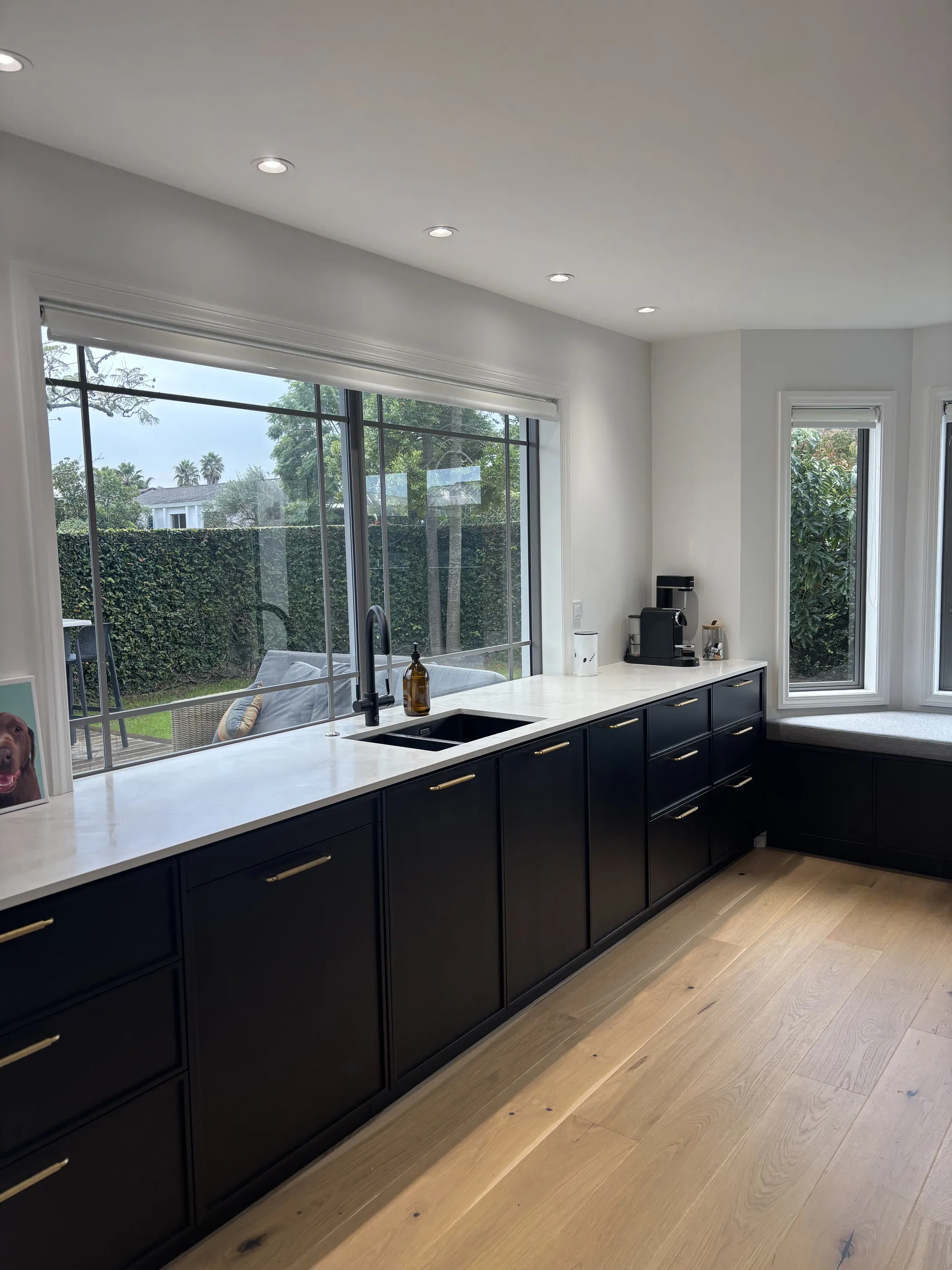 A run of black kitchen cabinetry with a white stone benchtop and brass handles, below a long window looking into the garden.