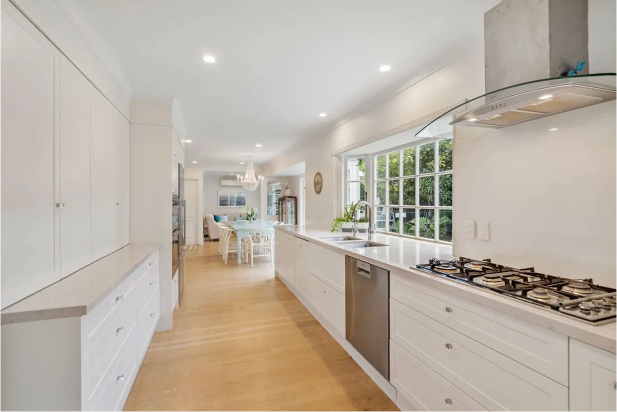 A long white galley kitchen with shaker cabinetry, stone benchtops, a garden window above the sink and a glass extractor over the cooktop.