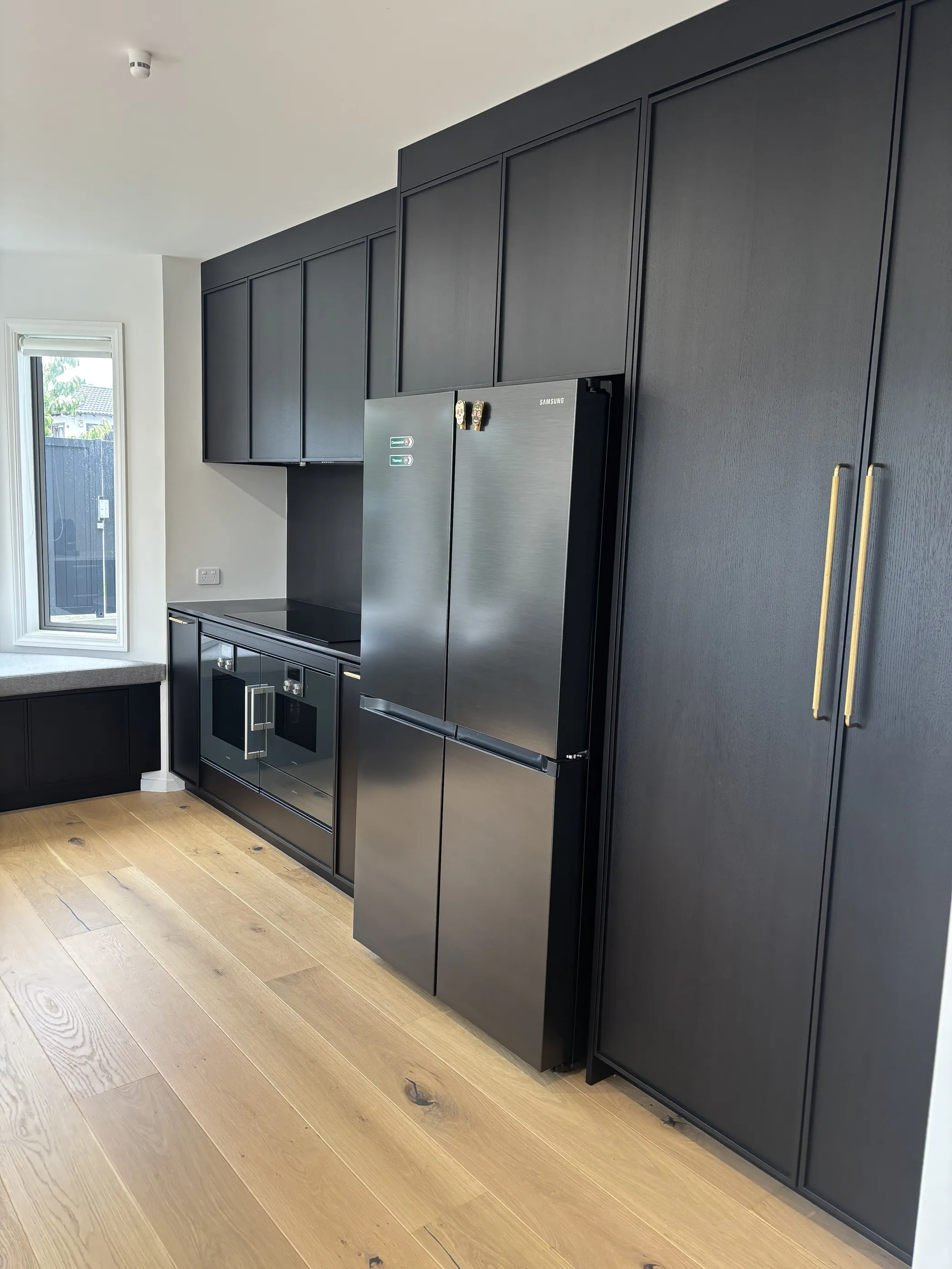 A kitchen with full-height black stained cabinetry and brass handles, integrated appliances and pale oak flooring.