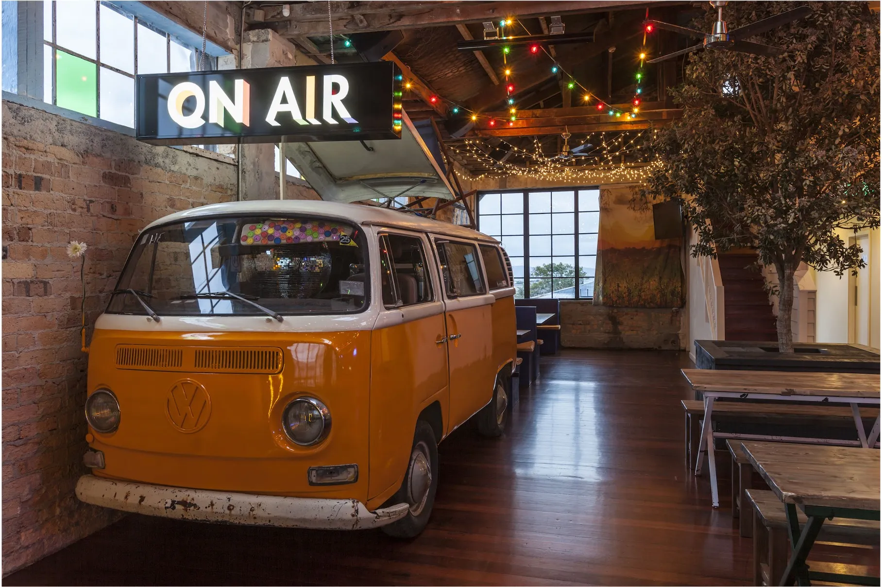 An orange Kombi van parked inside the venue below an illuminated "On Air" sign, with festoon lighting and timber floors around it.